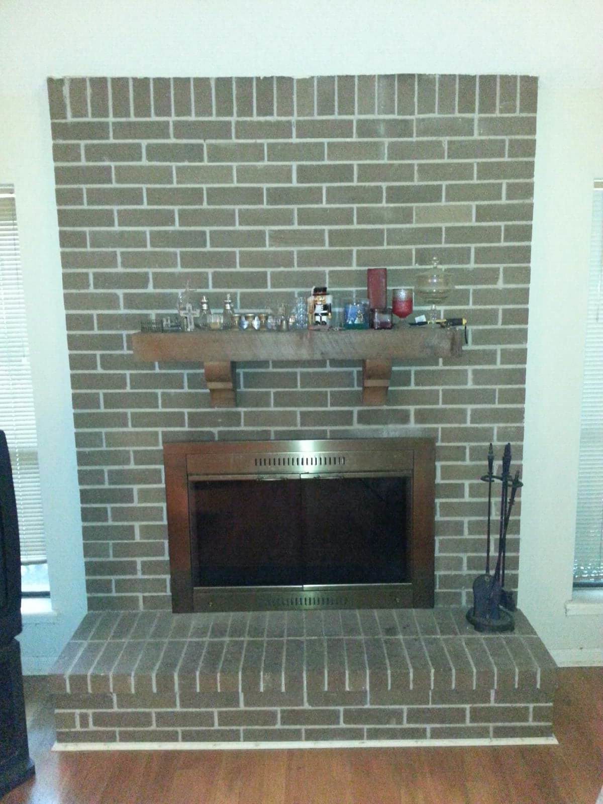 Large brick fireplace with large thick wood mantle decorated - front of brass fireplace and tools to the right.