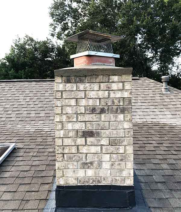Chimney covering liner on top of a chimney with colors of white, gray and black on composition roof.  Trees in background.
