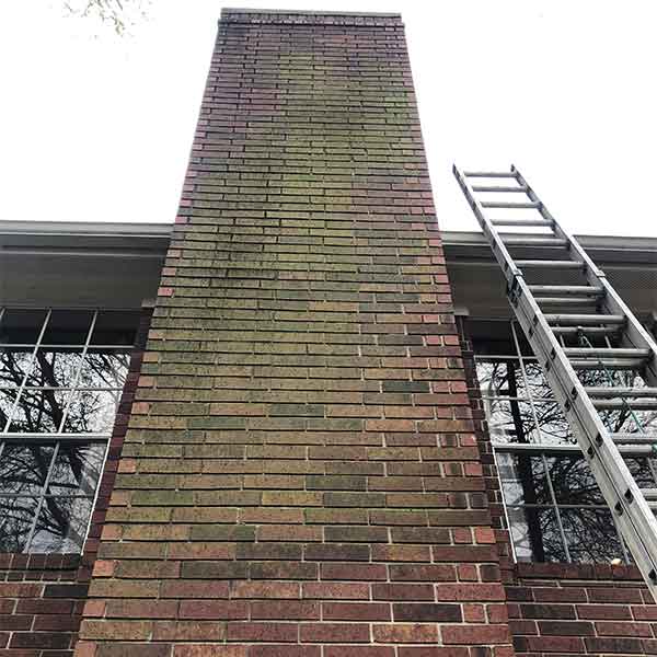 Very tall moldy brick fireplace before pressure wash with windows on each side and a ladder.