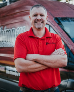 Anthony Kulhanek Technician in red logo shirt and short grayish hair.  Company truck with logo in background.
