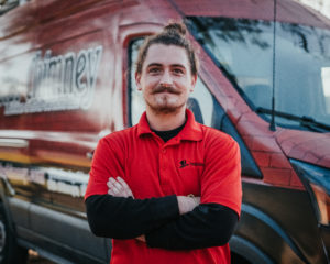 Michael Lyons technician with man bun and slight beard with logo shirt layered over black long sleeve.  Company truck with logo in background.